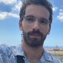Lion Blau, a man with dark curly hair, a beard and clear-framed glasses, in a blue patterned shirt under a blue sky with a city behind him.